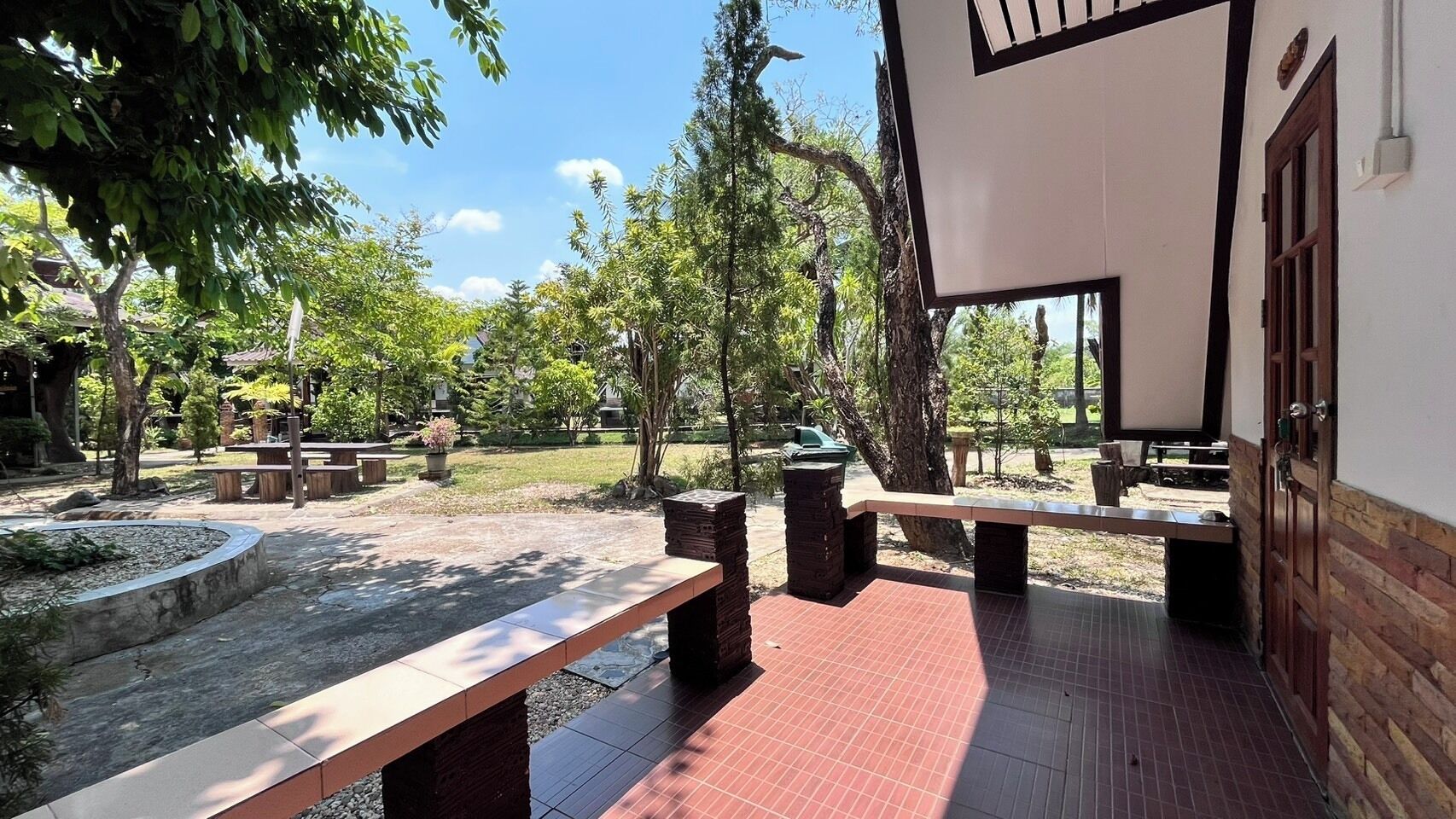 An outdoor patio area with benches and a tiled floor, surrounded by trees and greenery.