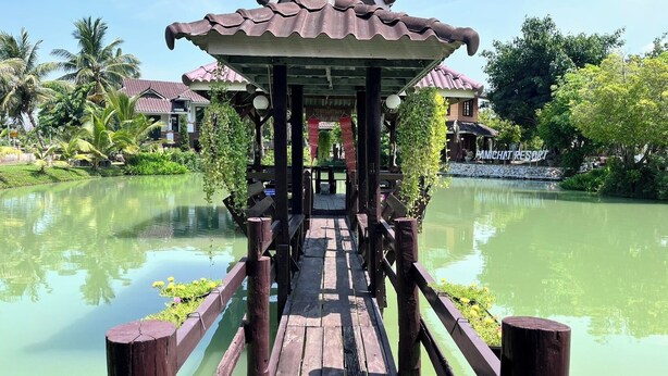 Un puente de madera que lleva a un edificio con techo de tejas, rodeado de agua y vegetación.