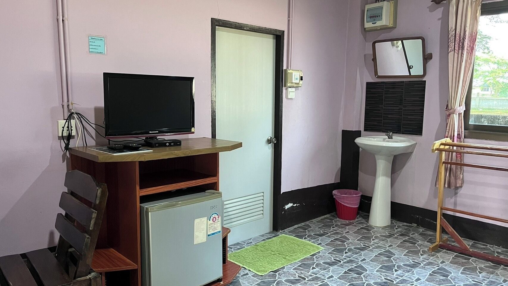 A small room with a television on a wooden stand, a sink, a mirror, and a window with curtains.
