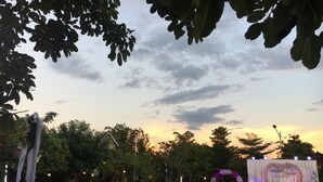 Un espacio para eventos al aire libre con mesas redondas, sillas y un escenario con adornos florales.