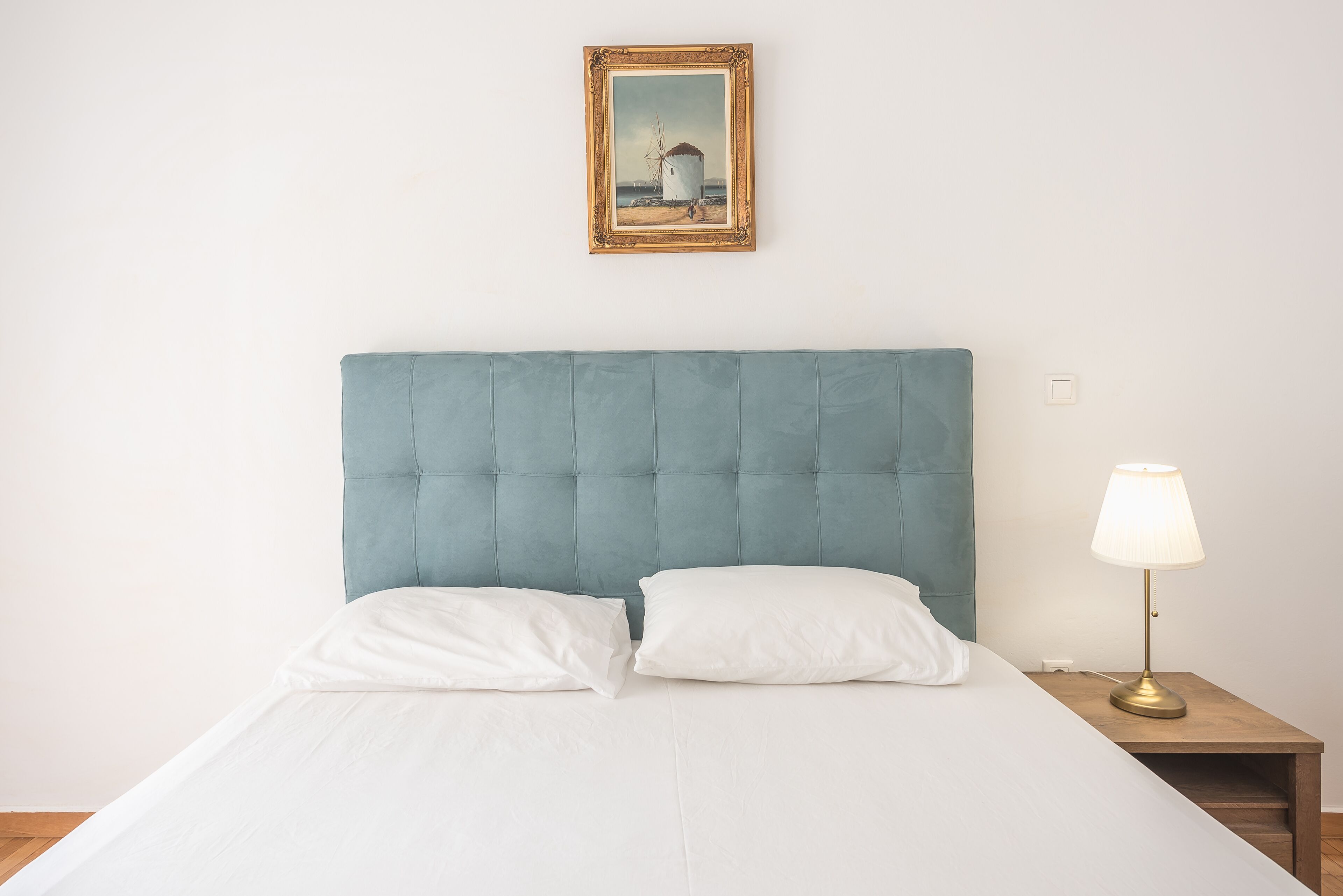 A neatly made bed with white linens, a blue upholstered headboard, and a bedside lamp on a wooden nightstand.