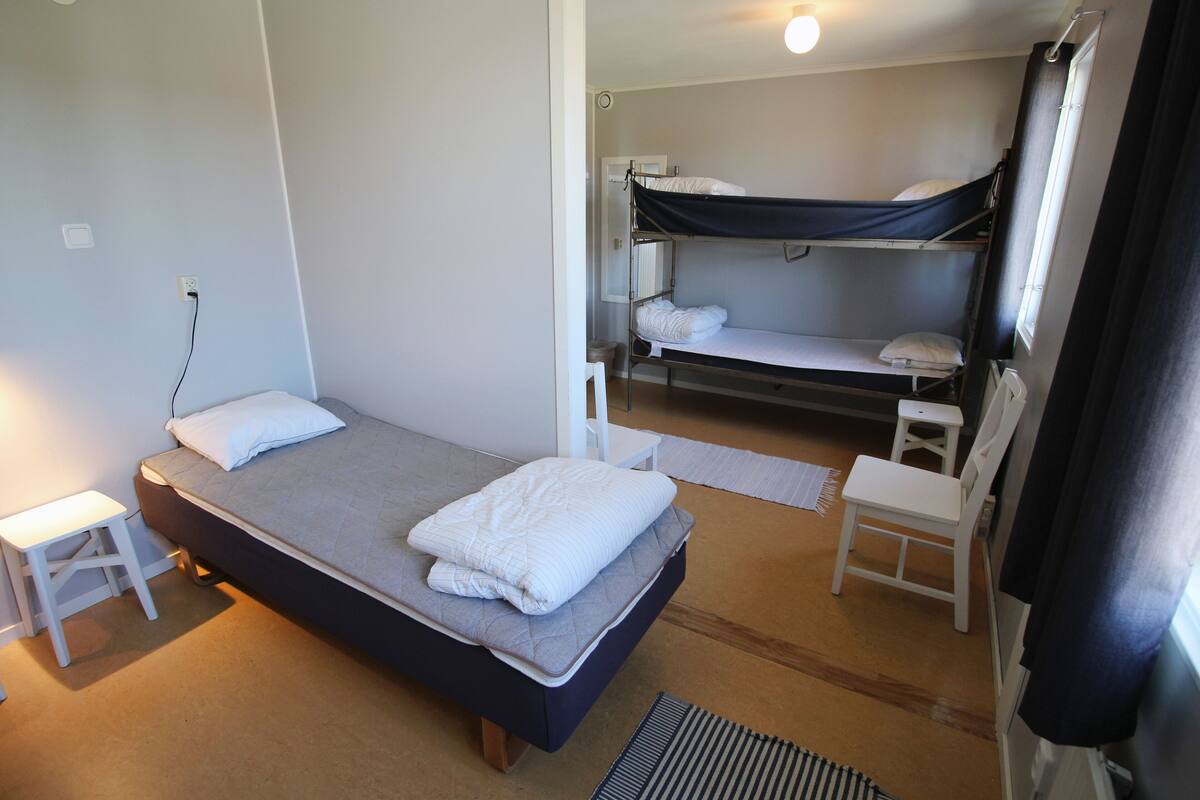 Ein kleines Zimmer mit einem Einzelbett, einem Etagenbett, einem kleinen weißen Stuhl und einem Nachttisch.