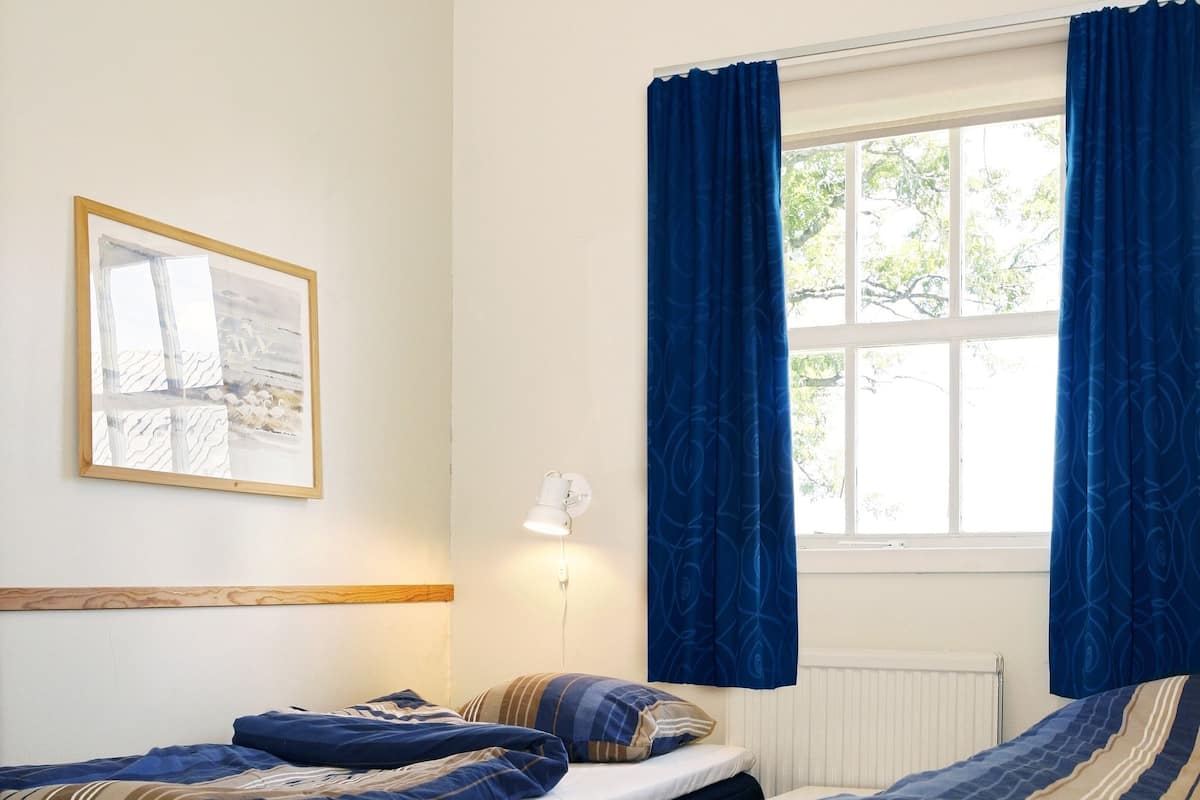 Ein Schlafzimmer mit zwei Betten, einem Fenster mit Vorhängen, einem Heizkörper und einem gerahmten Bild an der Wand.
