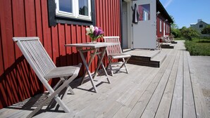 En träveranda med två vita stolar och ett bord, en röd byggnad med en vit dörr och en klarblå himmel.