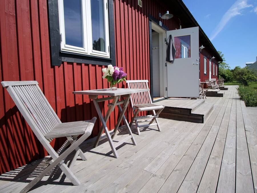 A wooden deck with two white chairs and a table, a red building with a white door, and a clear blue sky.