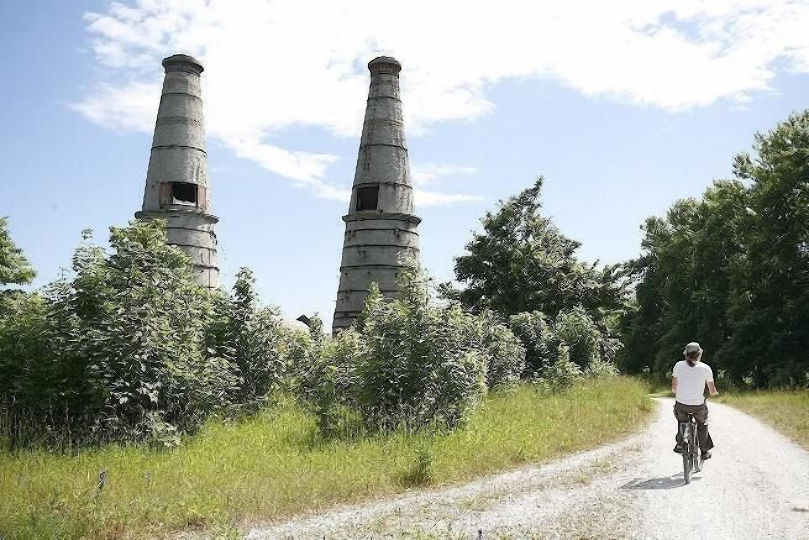 A person riding a bicycle on a dirt path with two large industrial chimneys in the background.