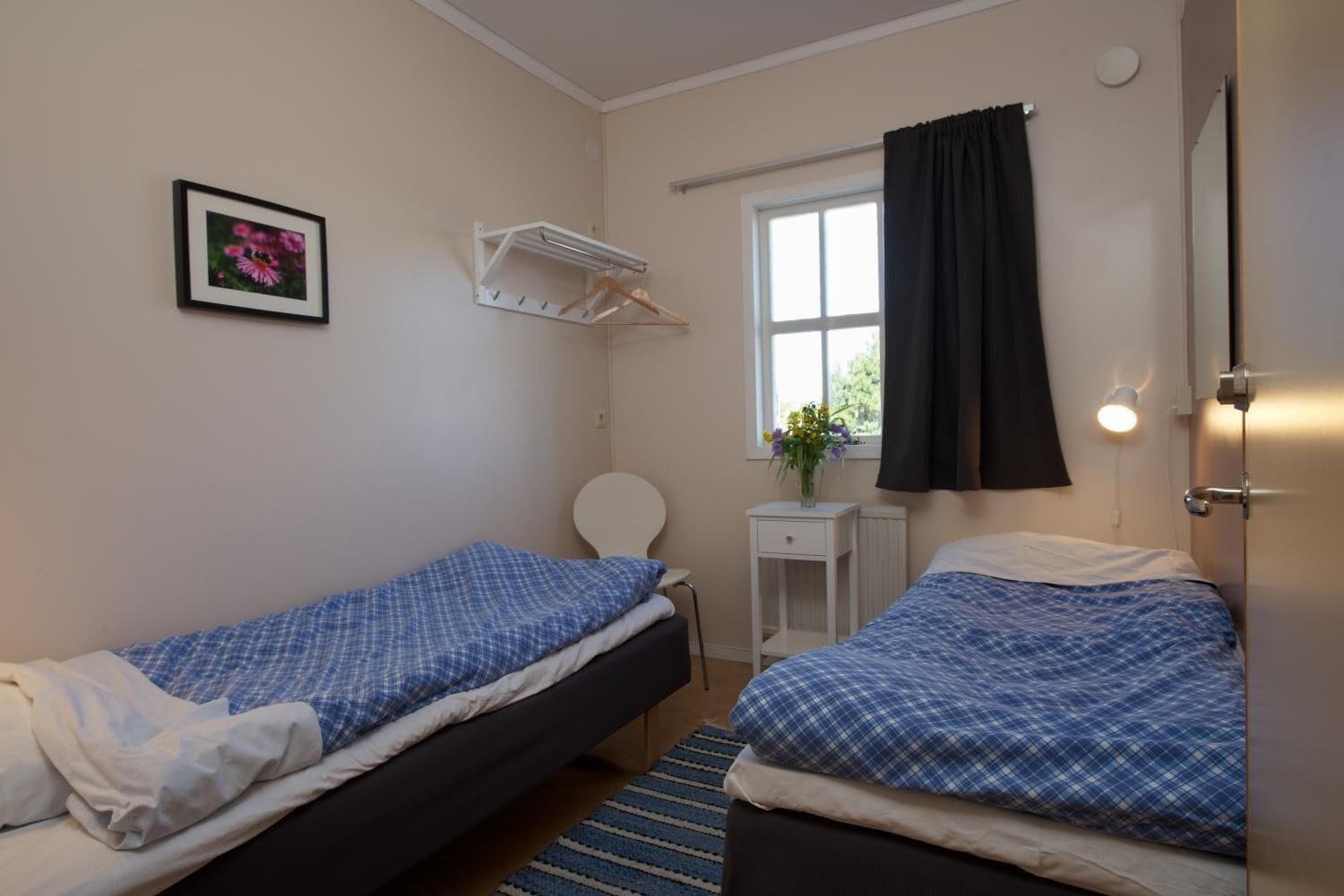 A small, clean room with two single beds, a white bedside table with a lamp, a window with a black curtain, and a framed picture on the wall.