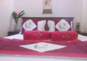 Cama arrumada com toalhas e almofadas vermelhas, um vaso com flores e uma obra de arte emoldurada na parede.
