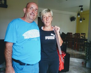 Um homem e uma mulher estão parados em um ambiente interno, com uma área de jantar ao fundo.