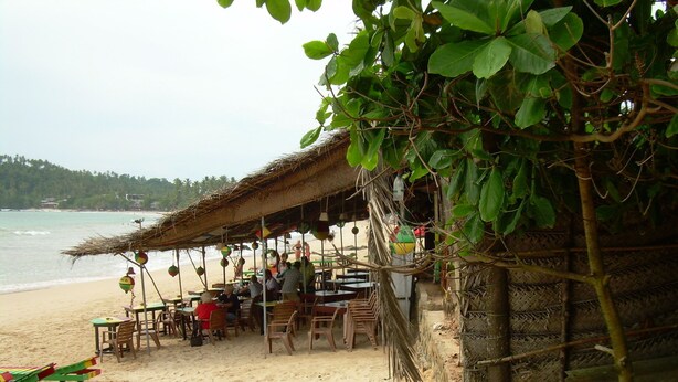 A beachside restaurant with outdoor seating, a thatched roof, and a view of the ocean. - Bandhusena Apartments (Mirissa)