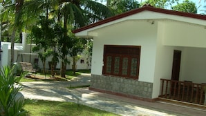 A small, single-story building with a white exterior and a red roof. It features a wooden door with glass panels and a small porch with a wooden railing. - Bandhusena Apartments (Mirissa)