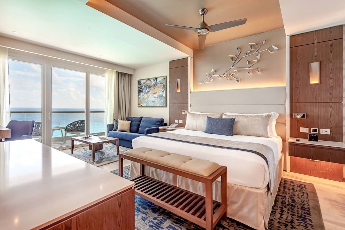 A modern hotel room with a large bed, a desk, a bench, and a view of the ocean.