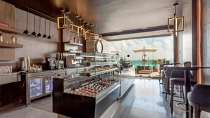 A modern café with a glass display case of pastries, a coffee bar, and a seating area with a view of the ocean. - Royalton CHIC Cancun, An Autograph Collection All-Inclusive Resort - Adults Only (Cancun)