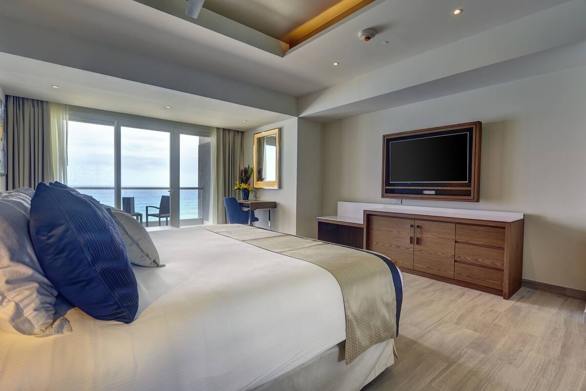 A modern hotel room with a large bed, a TV, a desk, and a view of the sea.