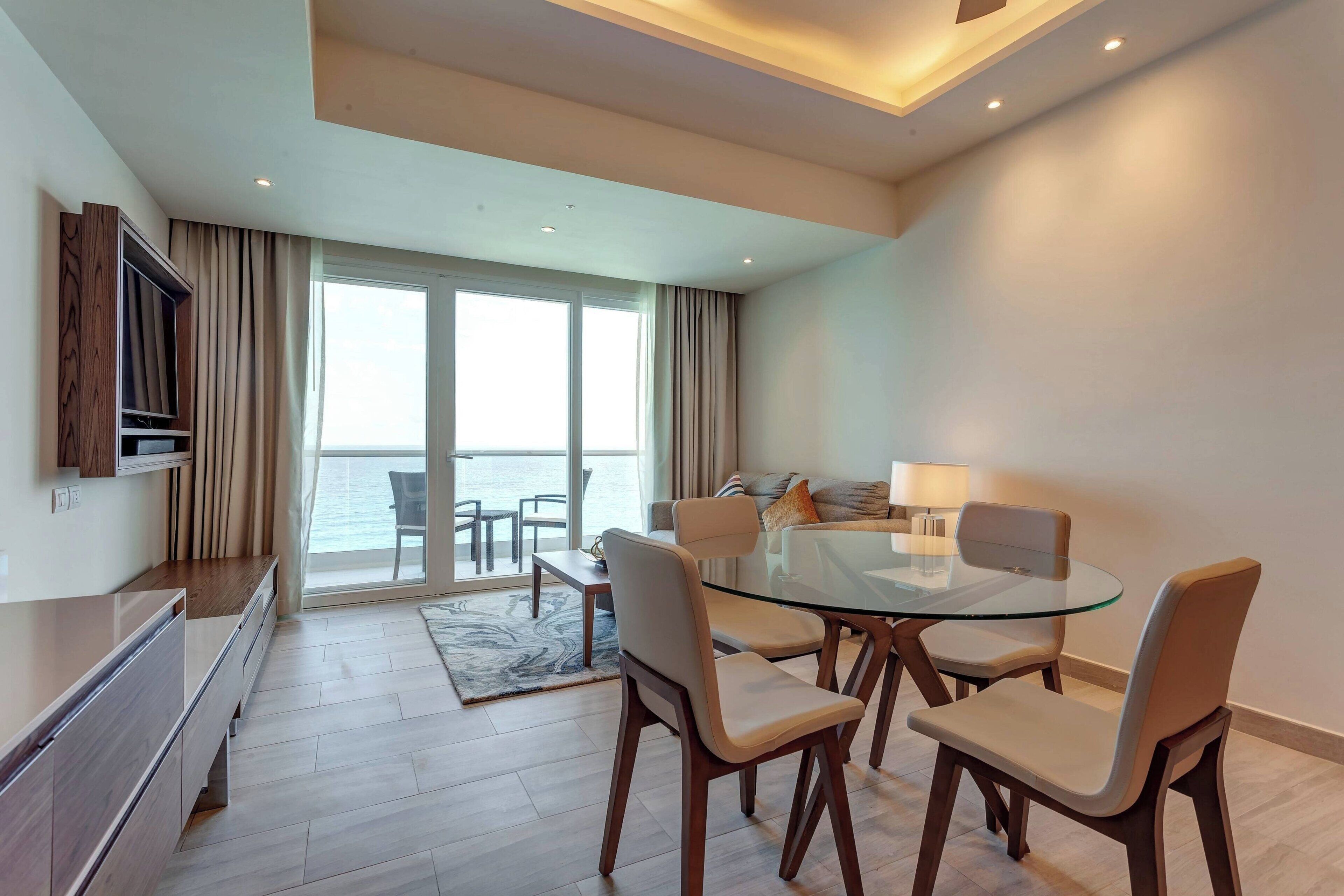 A modern living room with a glass dining table, wooden chairs, a sofa, and a TV mounted on the wall.