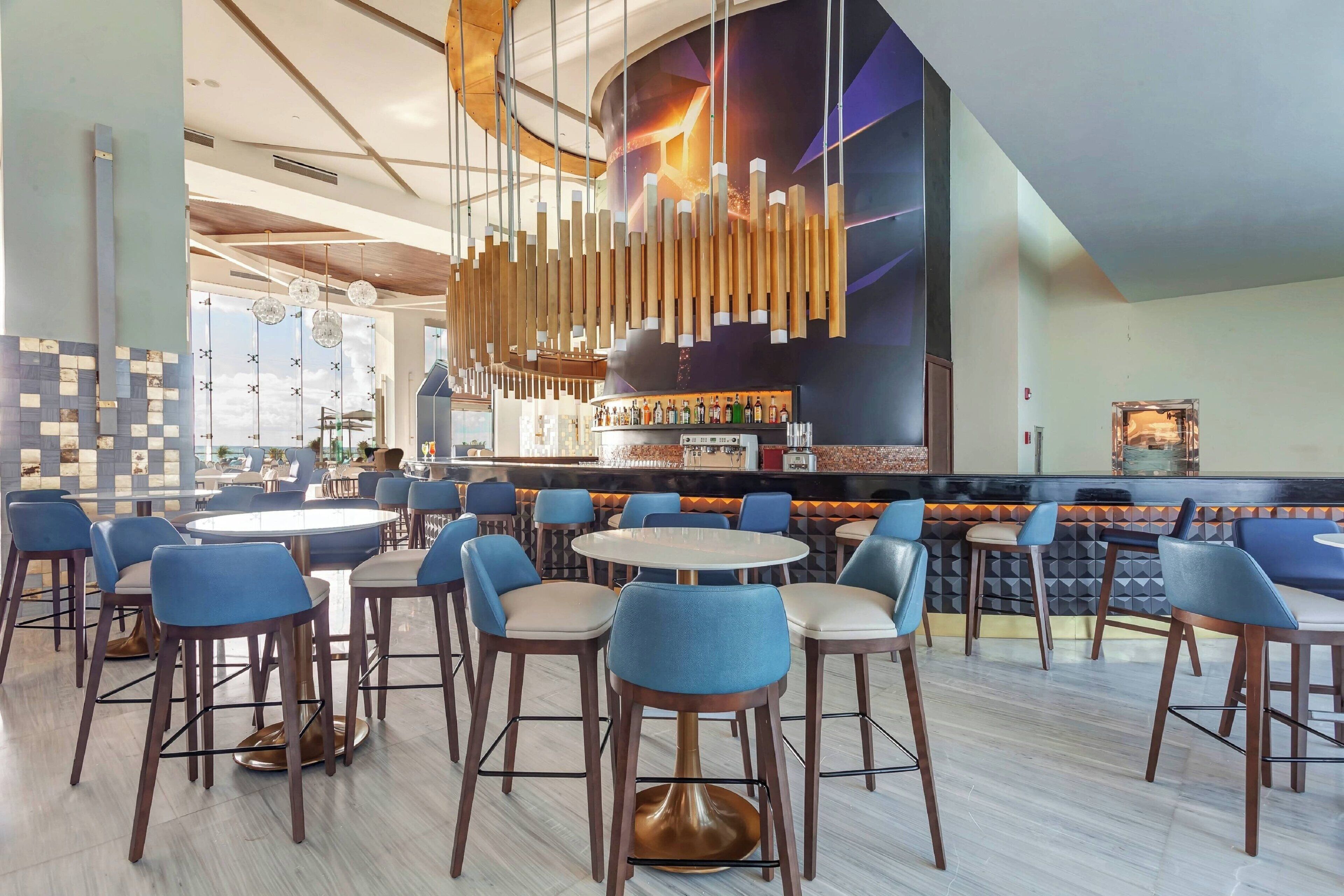 A modern restaurant with round tables and blue chairs, a bar area with a variety of bottles, and a large mural on the wall.