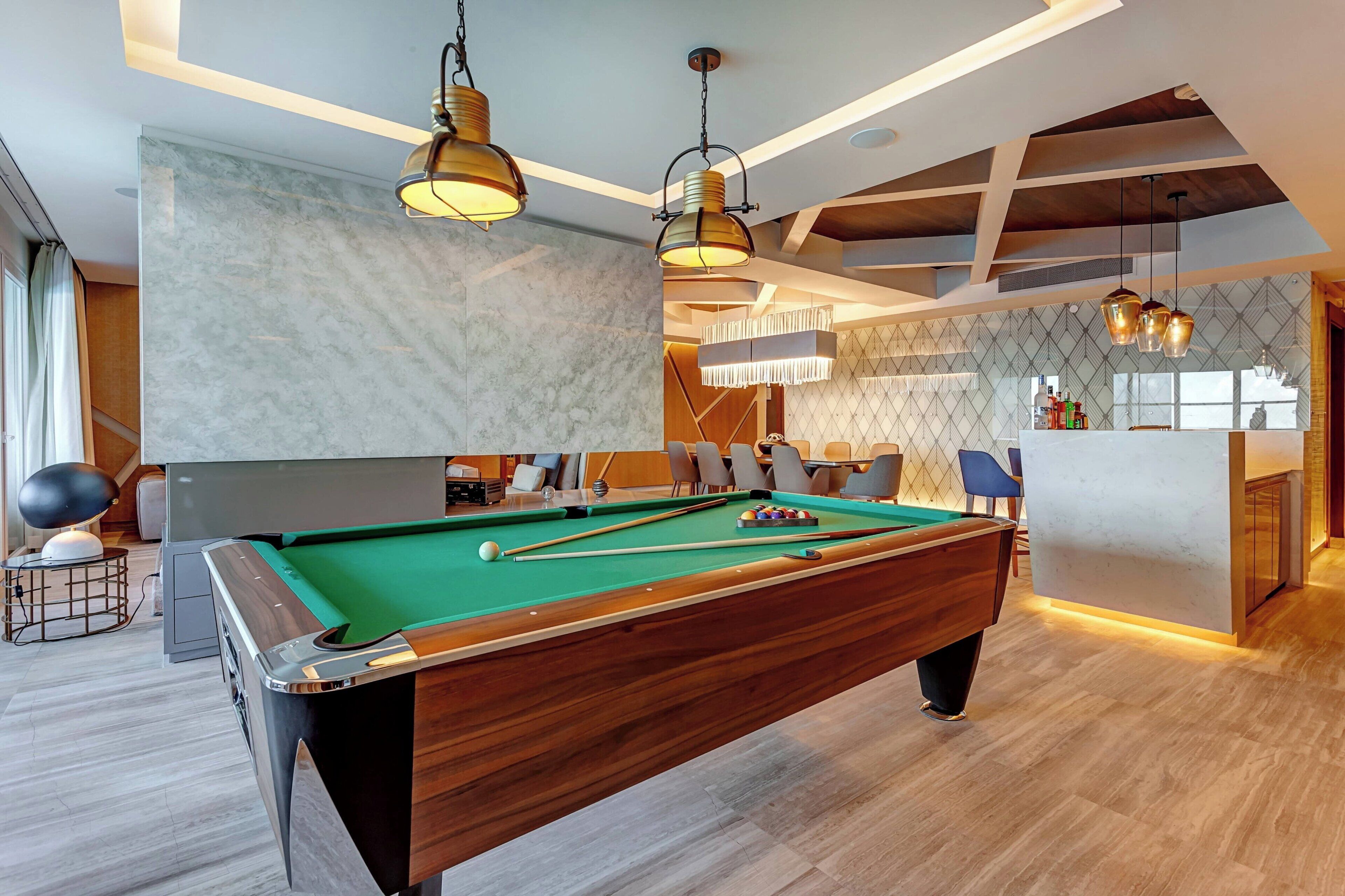 A modern pool table with a green felt surface, surrounded by wooden furniture and a bar area with a marble backsplash.