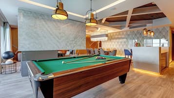 A modern pool table with a green felt surface, surrounded by wooden furniture and a bar area with a marble backsplash.