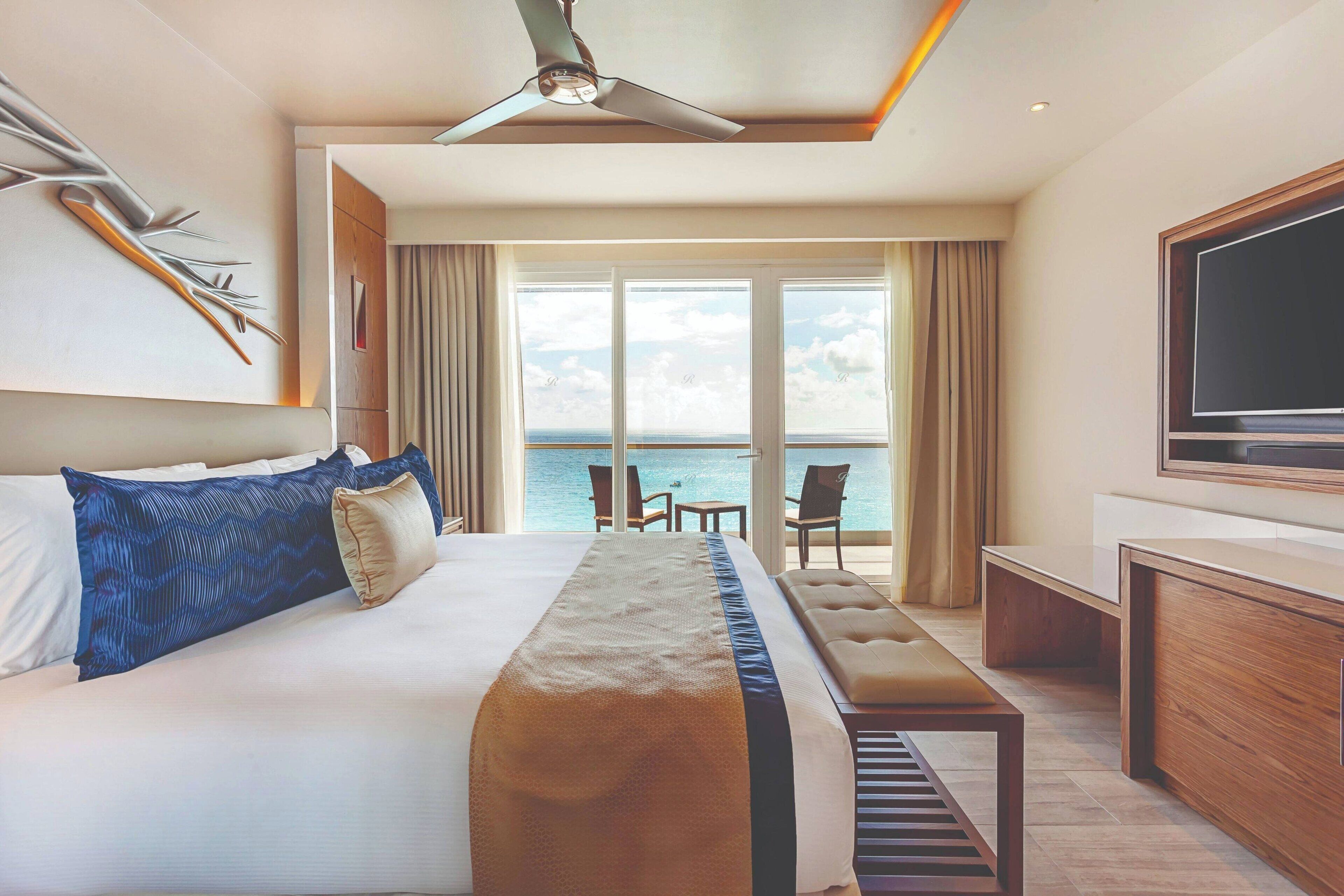 A hotel room with a large bed, a desk, a television, and a balcony with a view of the sea.