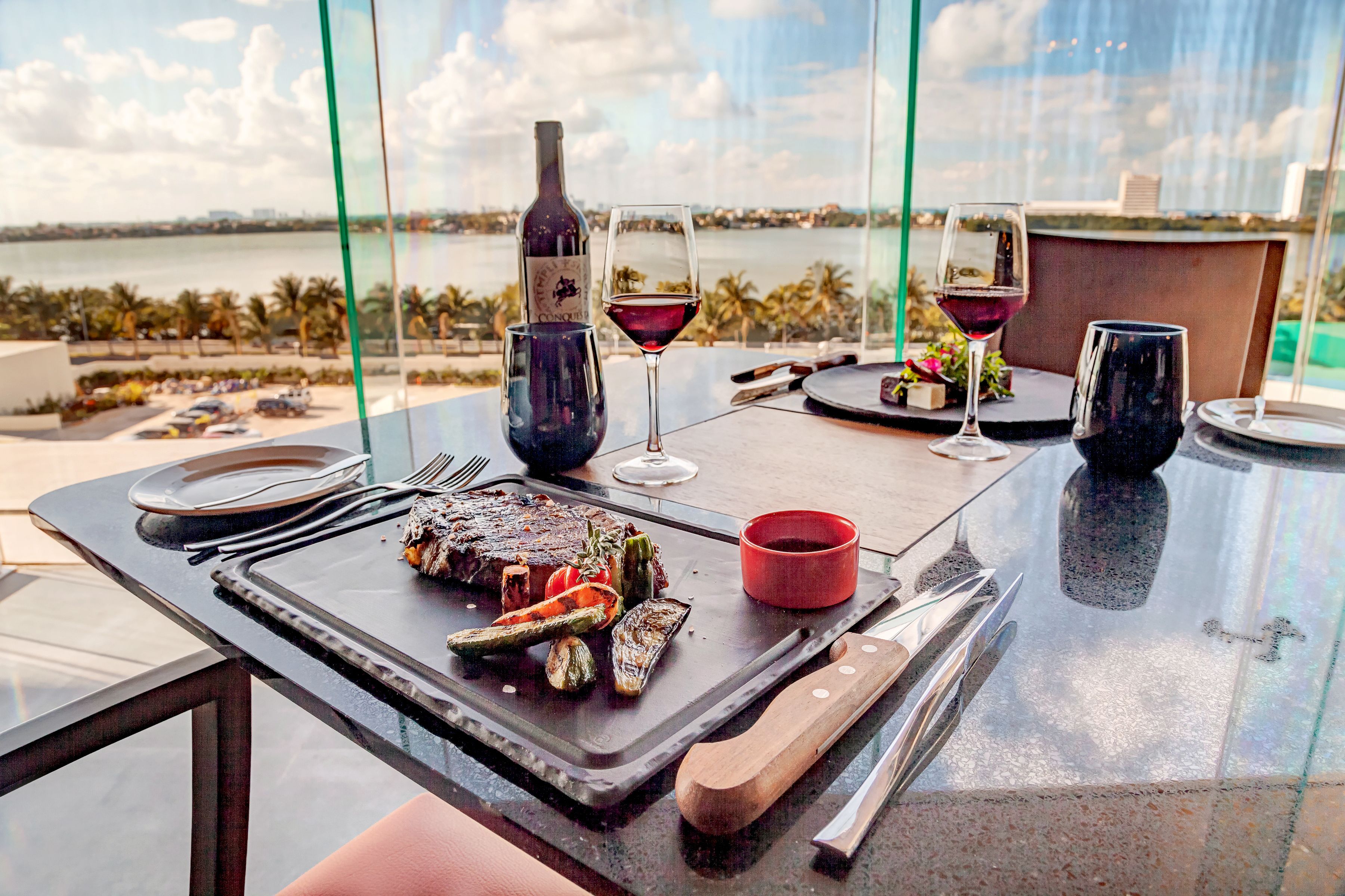 A dining table with a steak, vegetables, and wine glasses, overlooking a body of water.