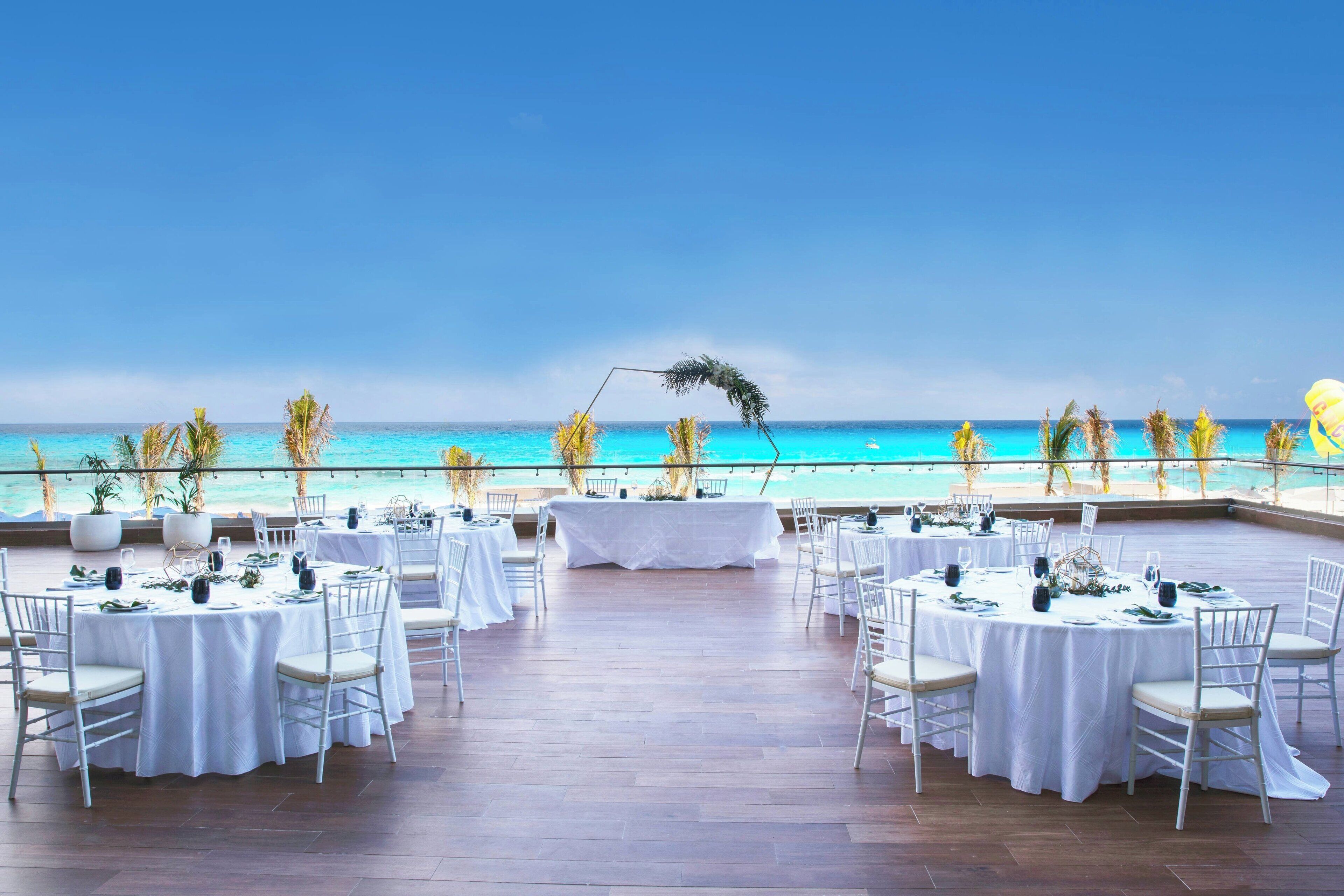 An outdoor dining area with round tables set for a wedding, overlooking a beach with palm trees and a clear blue sky.