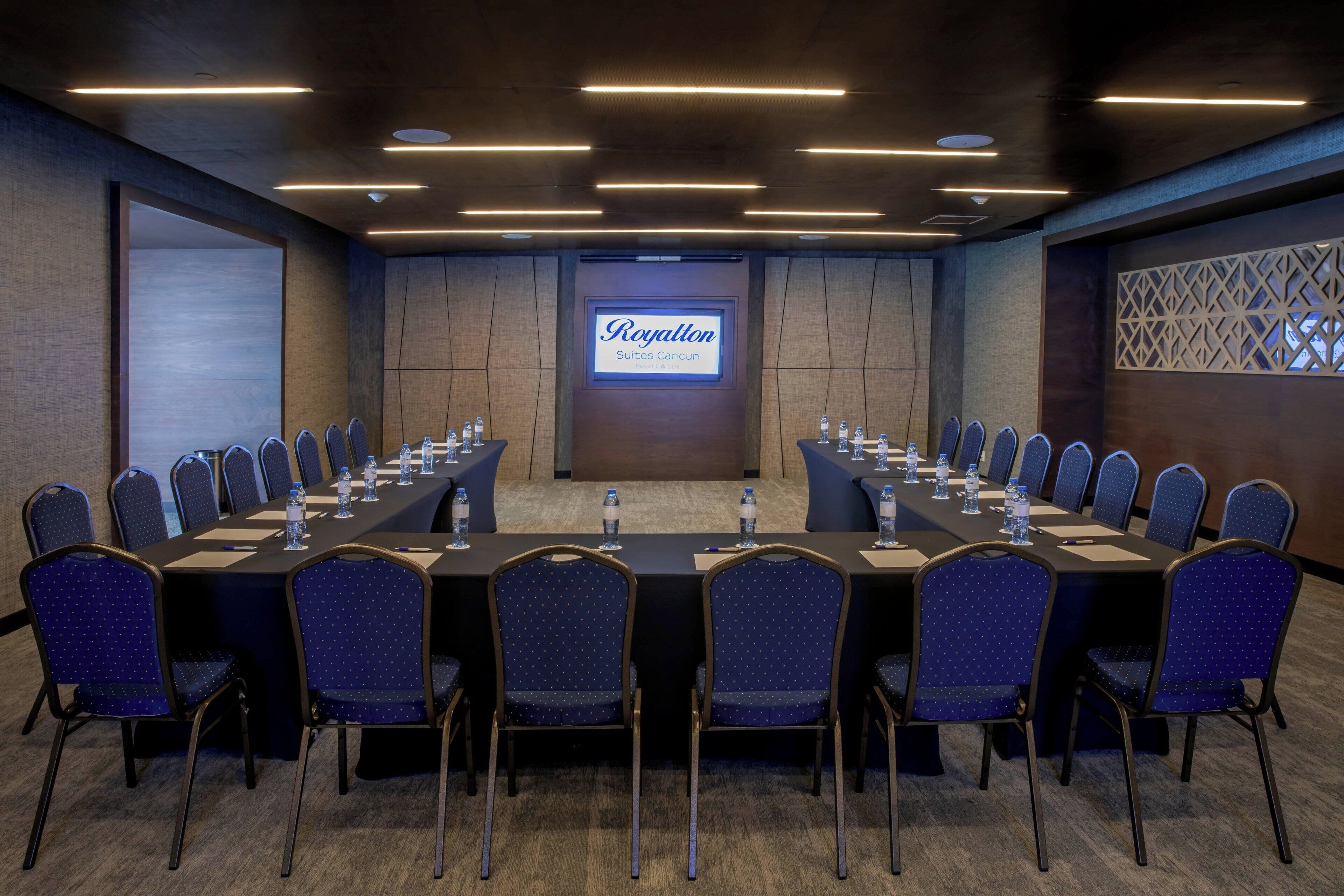 Un salón de conferencias con una larga mesa, sillas y una pantalla que muestra 'Royallton Suites Cancun'.