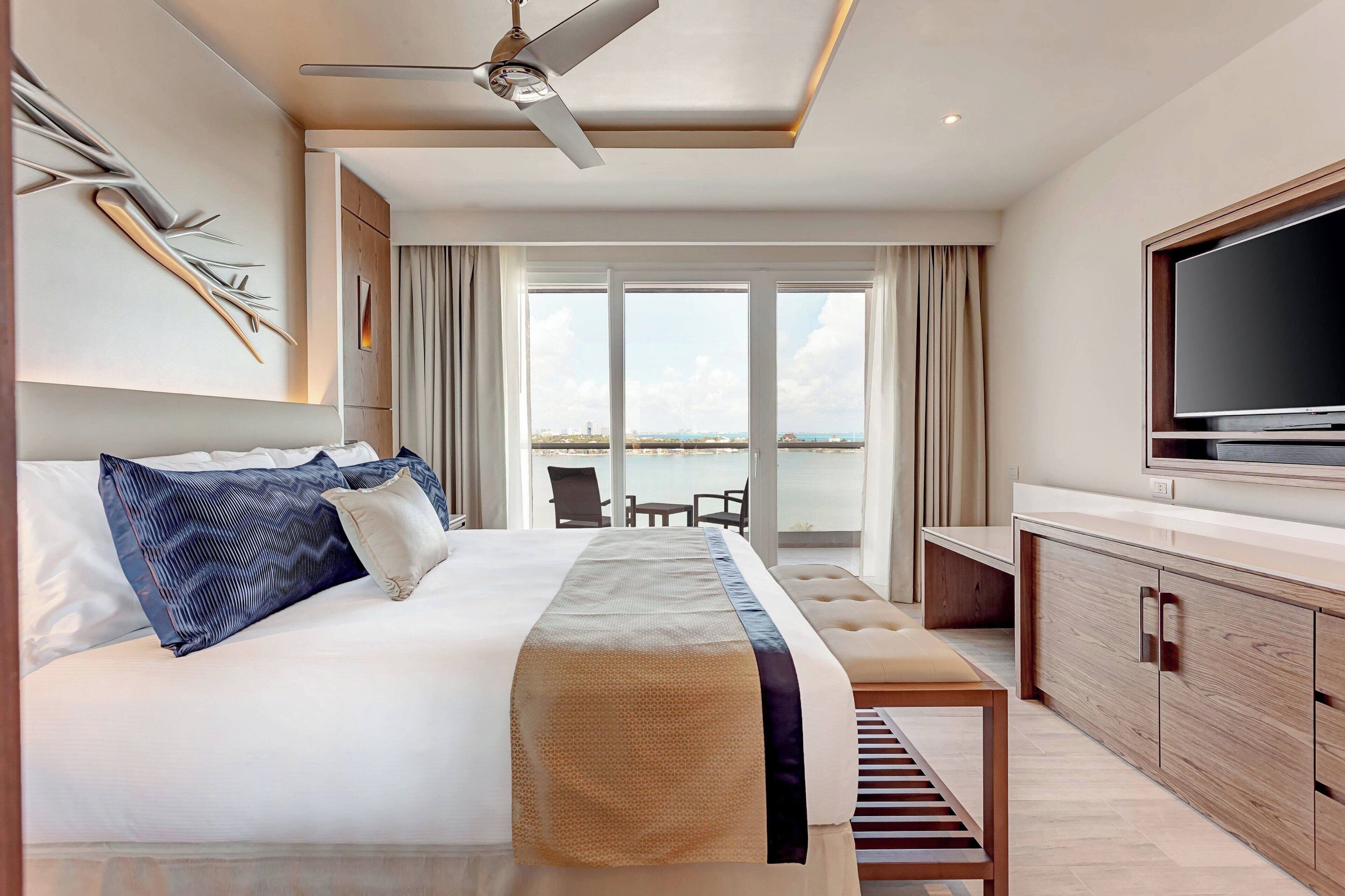 A modern hotel room with a bed, a TV, a ceiling fan, and a view of the sea.
