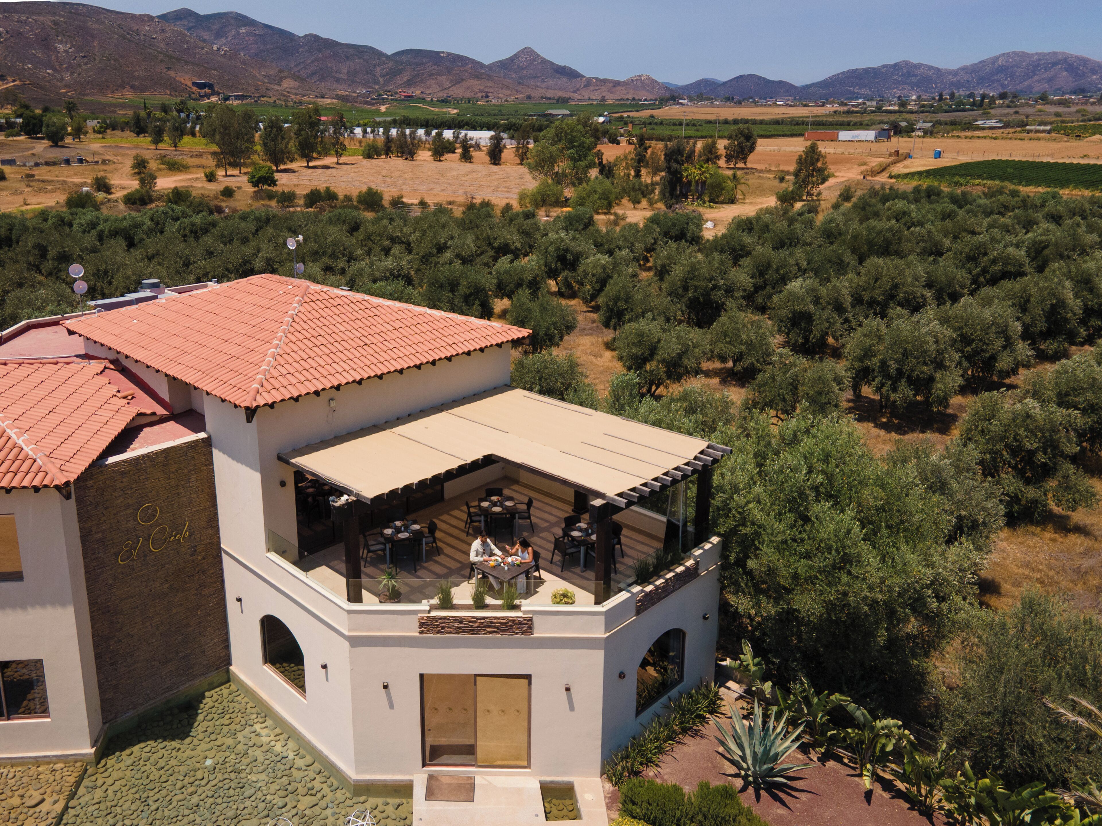 Edificio de dos plantas con tejado de tejas rojas, zona de estar al aire libre y vistas a olivares y montañas.