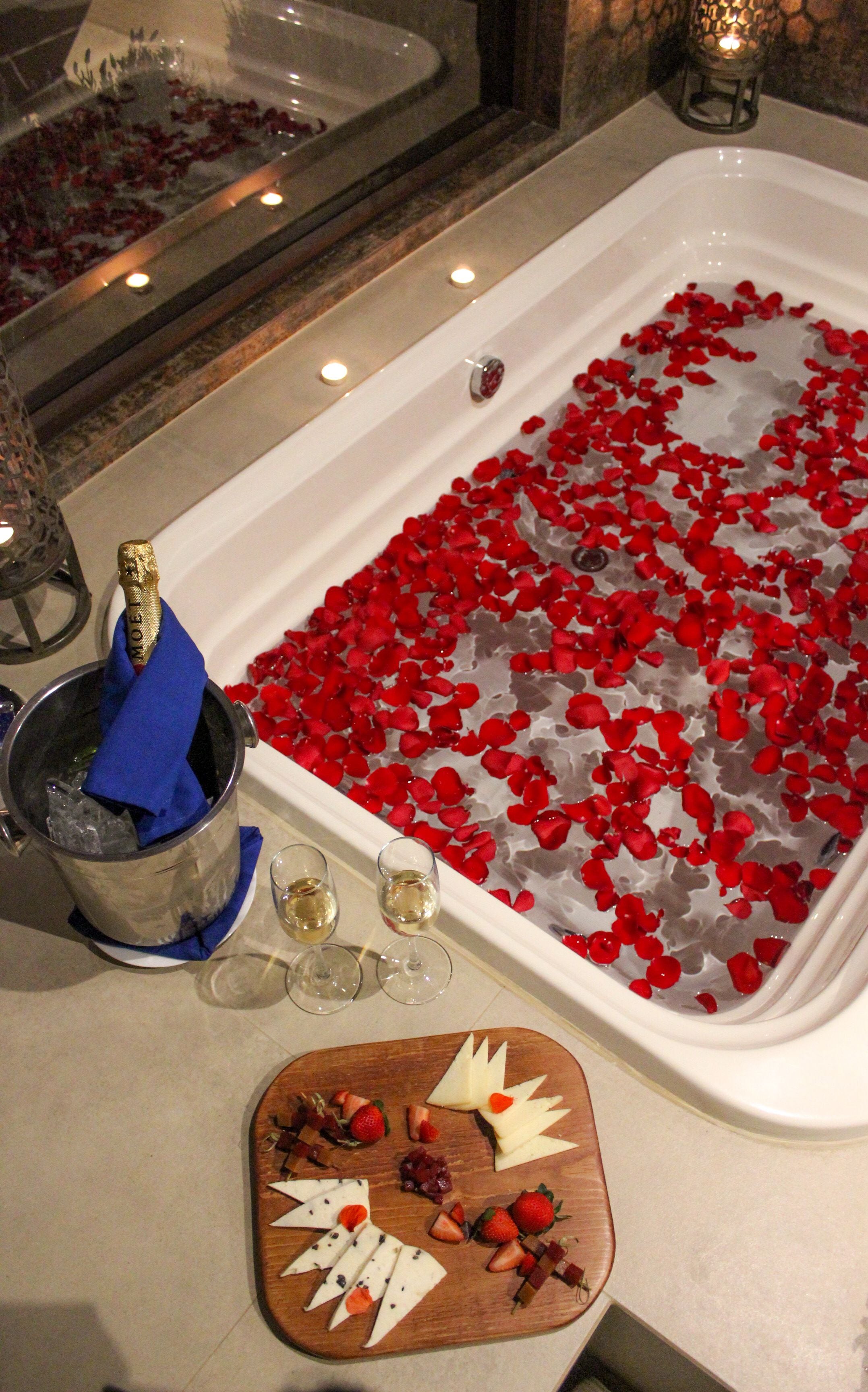 Una bañera llena de pétalos de rosa, una botella de champán y una tabla de quesos con fresas y uvas.