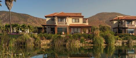 A two-story house with a balcony, a pond in the foreground, and hills in the background.
