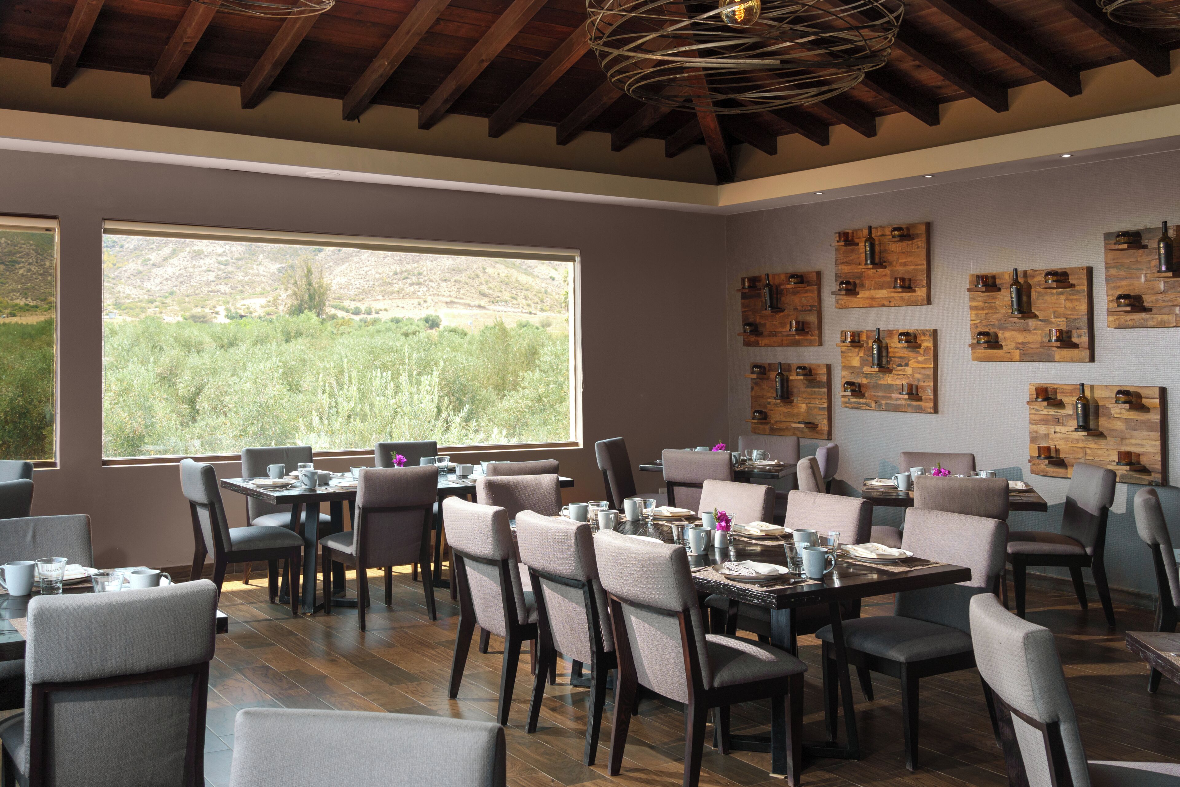 Un comedor con varias mesas y sillas, ventanas amplias con vistas a zonas verdes y decoración de pared de madera.