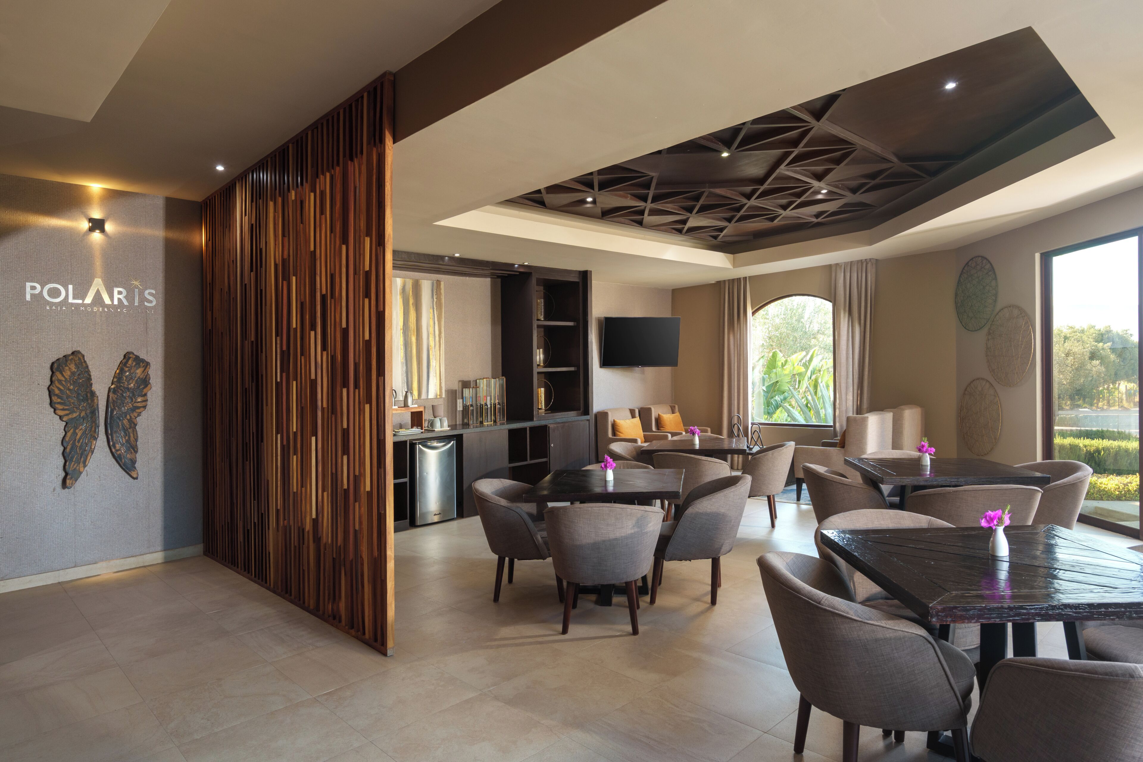 A modern hotel lobby with a dining area, a reception desk, and a large window offering a view of greenery.
