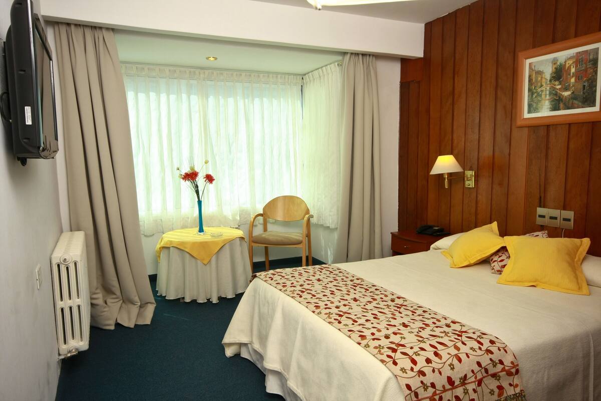 Una habitación de hotel con cama, una silla, una mesa con flores, una lámpara y un cuadro en la pared.
