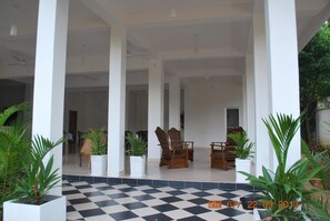 A covered outdoor area with a checkered floor, potted plants, wooden chairs, and a view into a building. - Dad's Holiday Home (Matale)