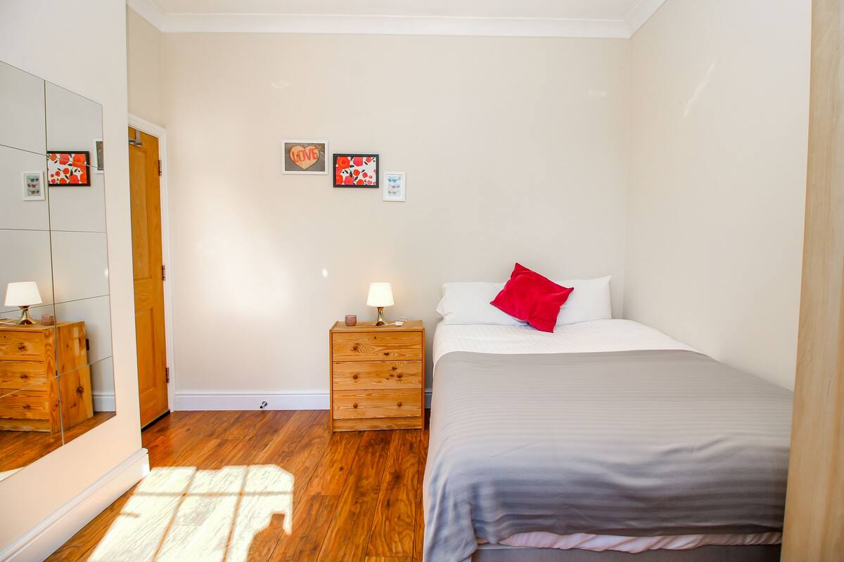A bedroom with a bed, a nightstand with a lamp, a dresser, and a mirror.