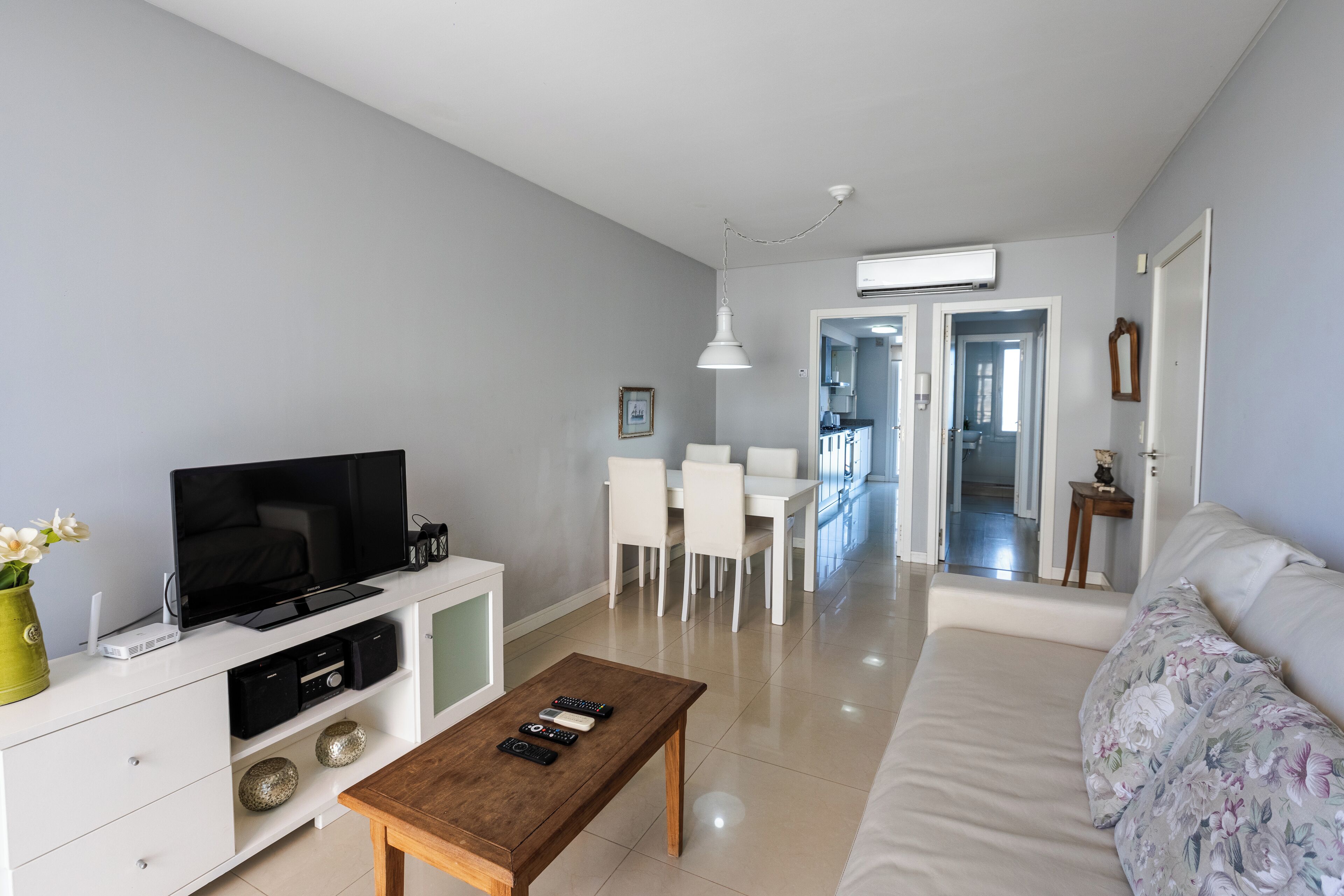 Ein modernes Wohnzimmer mit Fernseher, Couchtisch, Sofa und einem Essbereich mit weißen Stühlen und einem Tisch.