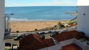 A beach view from a building with a tiled roof, a road with parked cars, and a clear sky. - DeptosVip (Mar del Plata)