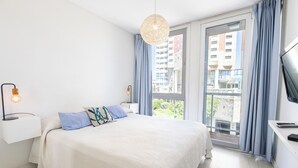 A modern bedroom with a large bed, a bedside table with a lamp, a TV, and a view of buildings through a window. - DeptosVip (Mar del Plata)