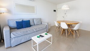 A modern living room with a grey sofa, a small white table with two decorative items, and a wooden dining table with white chairs. - DeptosVip (Mar del Plata)