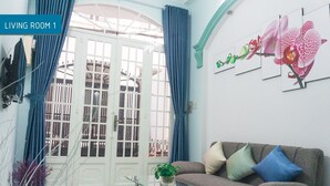 Un salón con una mesa de centro de vidrio, un sofá con cojines, una puerta con cortinas y una obra de arte en la pared con orquídeas.