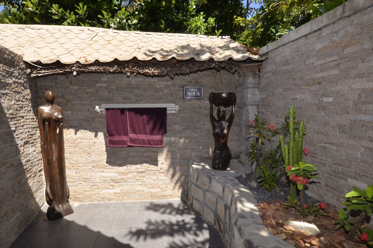 Un petit bâtiment aux murs de pierre, surmonté d’un toit en tuiles, abritant une statue en bois, une enseigne suspendue portant l’inscription « Chez Nous », et un petit jardin agrémenté de cactus et de fleurs.