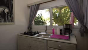 A compact kitchen with a gas stove, sink, and a window with curtains. - Le Laugaut's Bungalows (Nosy Be)