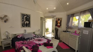 A bedroom with a large bed, a bedside table, a nightstand, a fan, and a window with curtains. - Le Laugaut's Bungalows (Nosy Be)