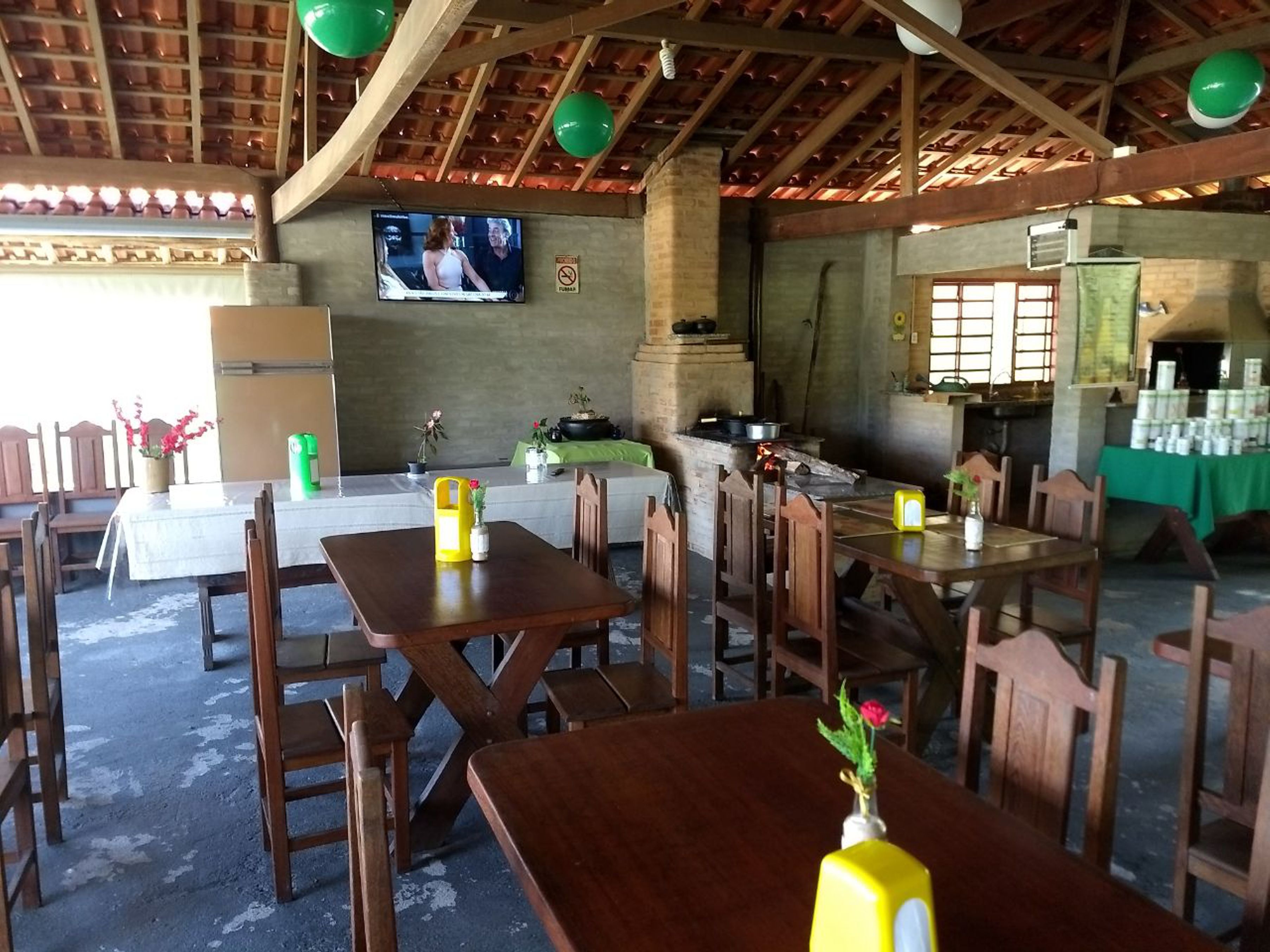 A rustic dining area with wooden tables and chairs, a television mounted on the wall, and a brick oven in the background.