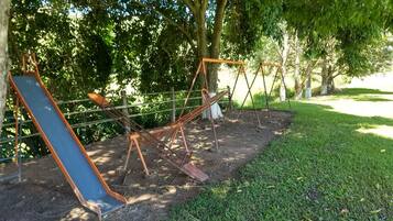 A playground with swings and a slide, surrounded by trees and grass.