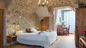 A bedroom with a stone wall, a wooden door, a bed with white linens, a chandelier, and a view of an outdoor patio. - Can Barrull (Palau-Sator)