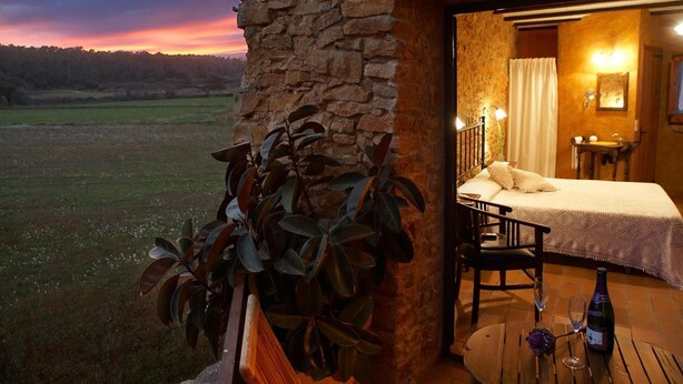 A stone building with a bedroom featuring a bed, bedside table, and a small table with a bottle and glasses. - Can Barrull (Palau-Sator)