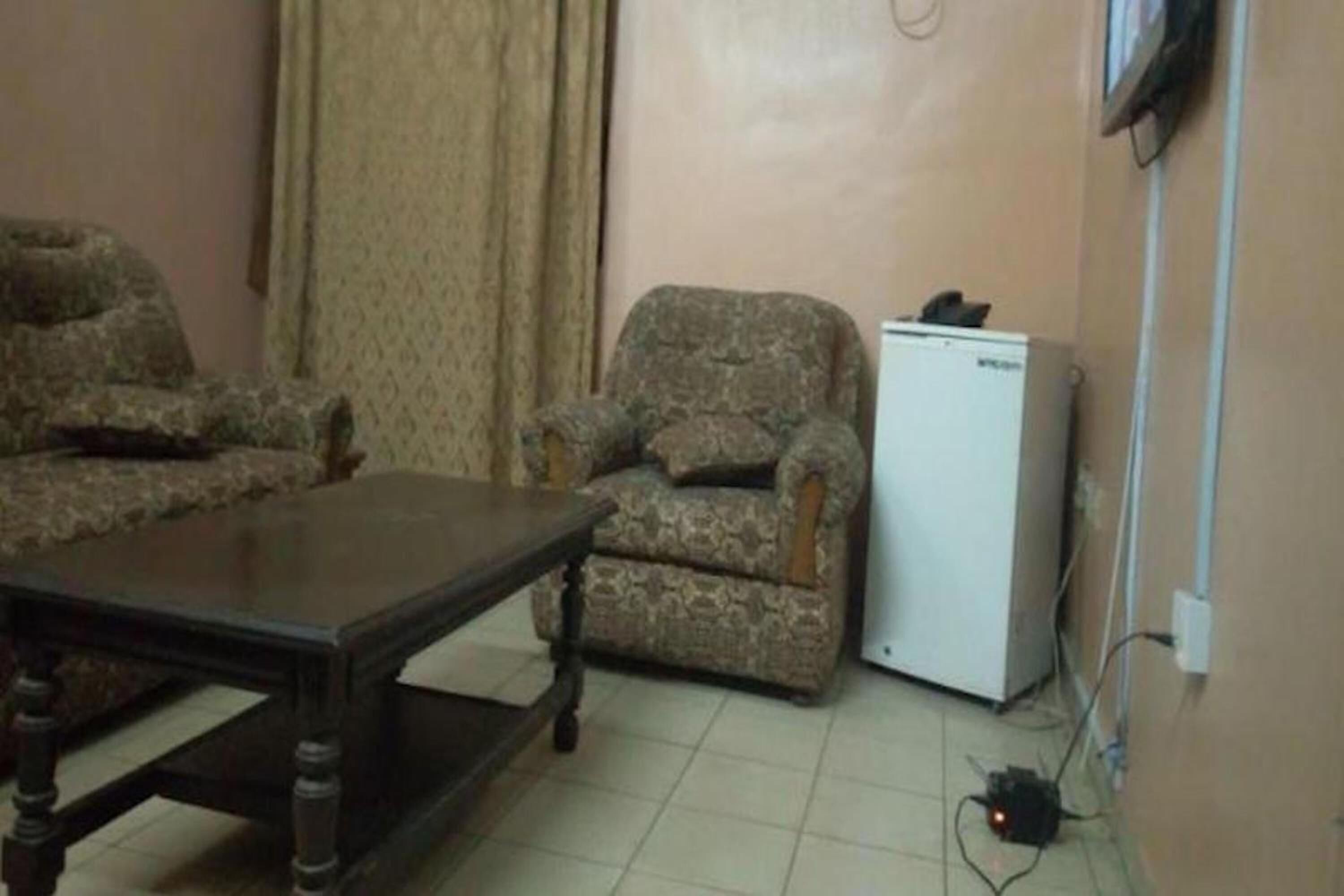 Una habitación con una mesa de madera oscura, dos sillones con estampado y un refrigerador blanco.