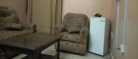 Una habitación con una mesa de madera oscura, dos sillones con estampado y un refrigerador blanco.