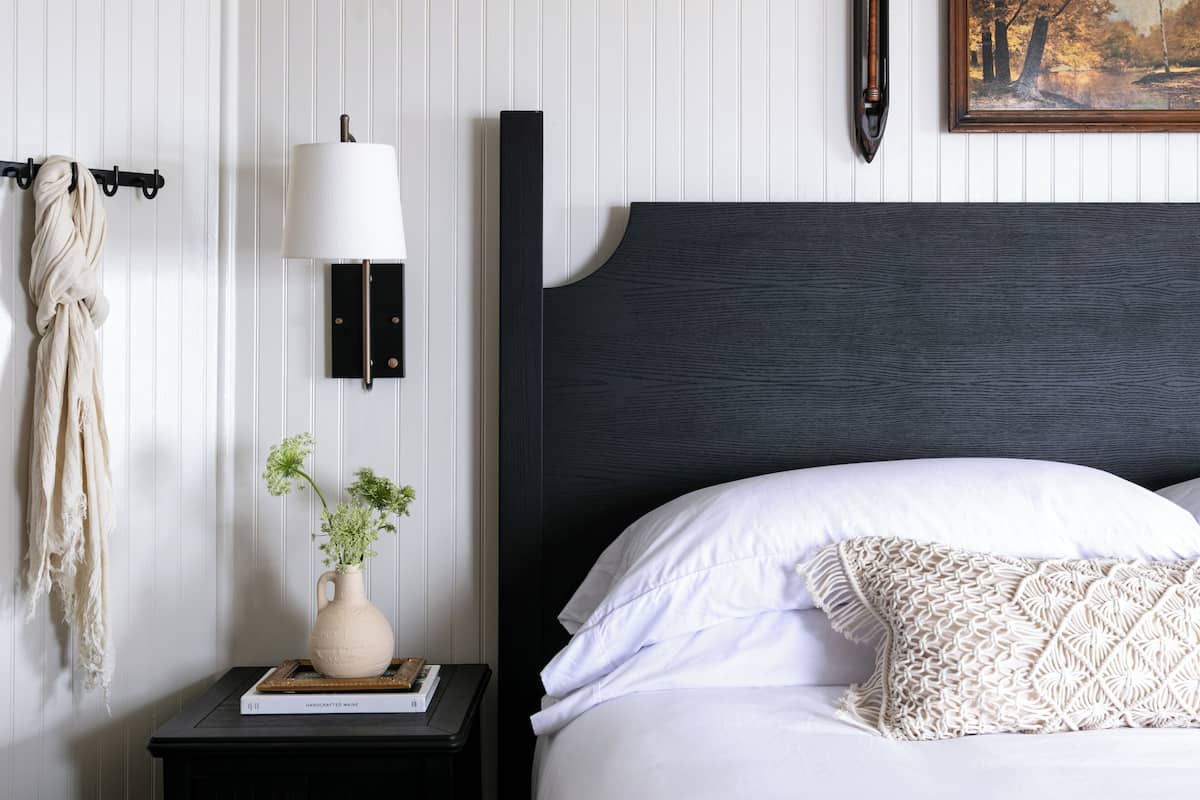 Ein Schlafzimmer mit dunkelbraunem Kopfteil, weißer Bettwäsche, einer Nachttischlampe, einem gerahmten Gemälde und einem kleinen Nachttisch mit einer Vase und einem Buch.